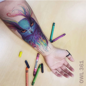 Woman's arm with owl temporary tattoo on top of a wooden table with crayons.